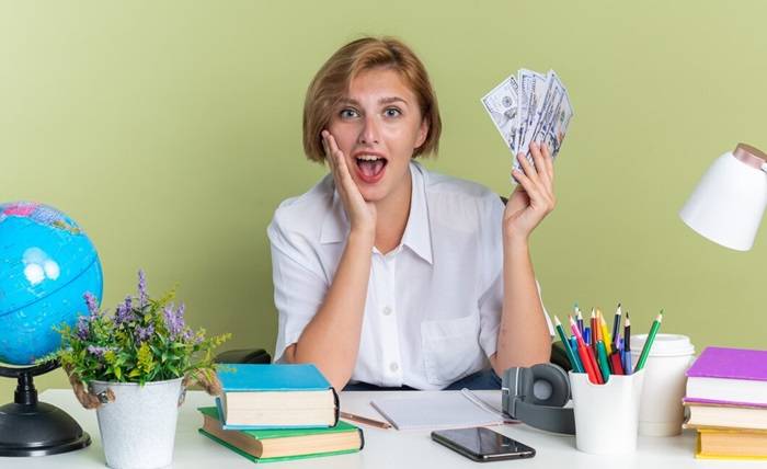 surprised young blonde student girl sitting desk with school tools keeping hand face holding money 141793 112082
